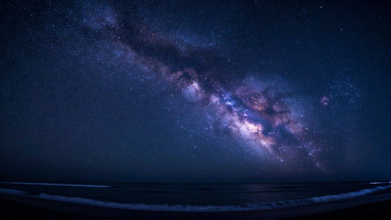 Cielo nocturno estrellado con nebulosas púrpuras sobre la silueta de olas en una costa tropical.