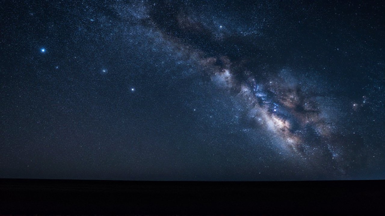 Vasto campo de estrellas brillantes y constelaciones sobre un paisaje terrestre oscuro bajo un cielo índigo.