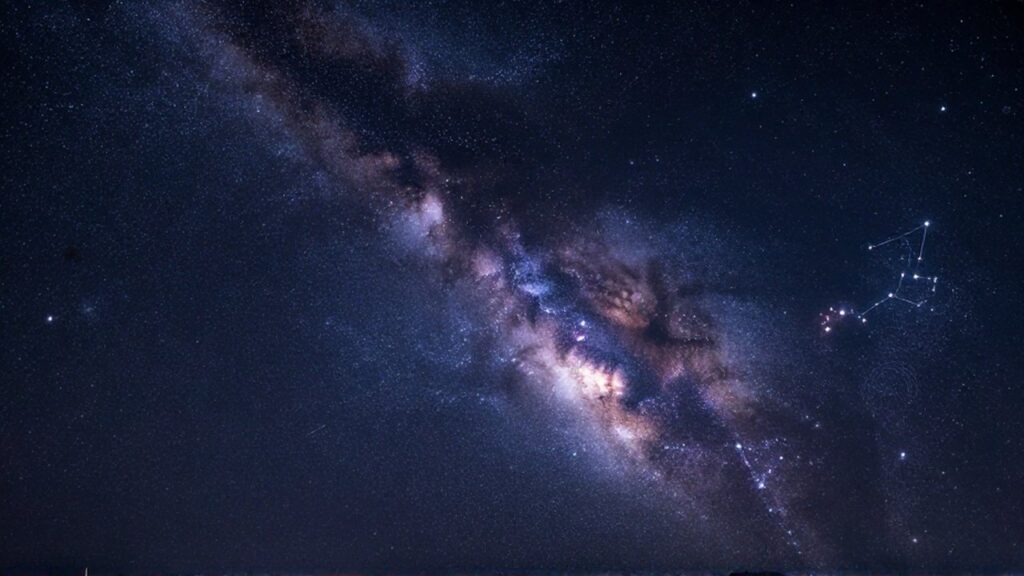 Vía Láctea brillante con nebulosas y cúmulos de estrellas sobre un cielo nocturno violeta.