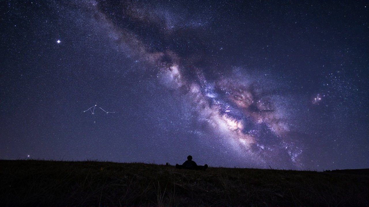 Persona recostada en una colina oscura contemplando la Vía Láctea y constelaciones en un cielo estrellado.